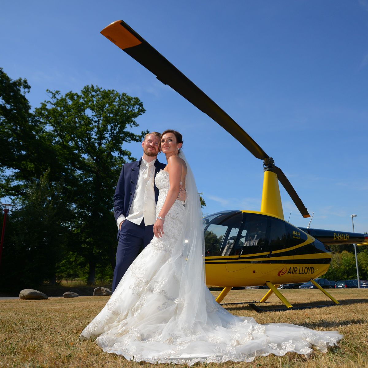 A bride and groom stand in front of a yellow AirLloyd helicopter.
