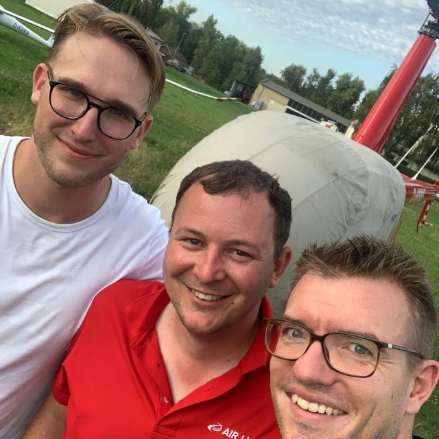 Drei Männer stehen vor einem roten Helikopter und lächeln in die Kamera.