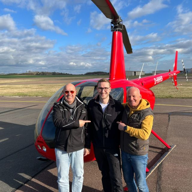 Drei Männer stehen vor einem roten Helikopter auf einem Fluglandeplatz.