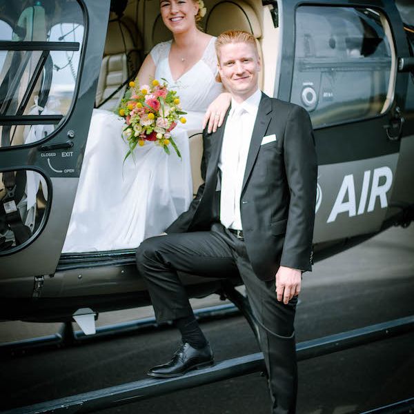 A bridal couple poses in front of a black AirLloyd helicopter.