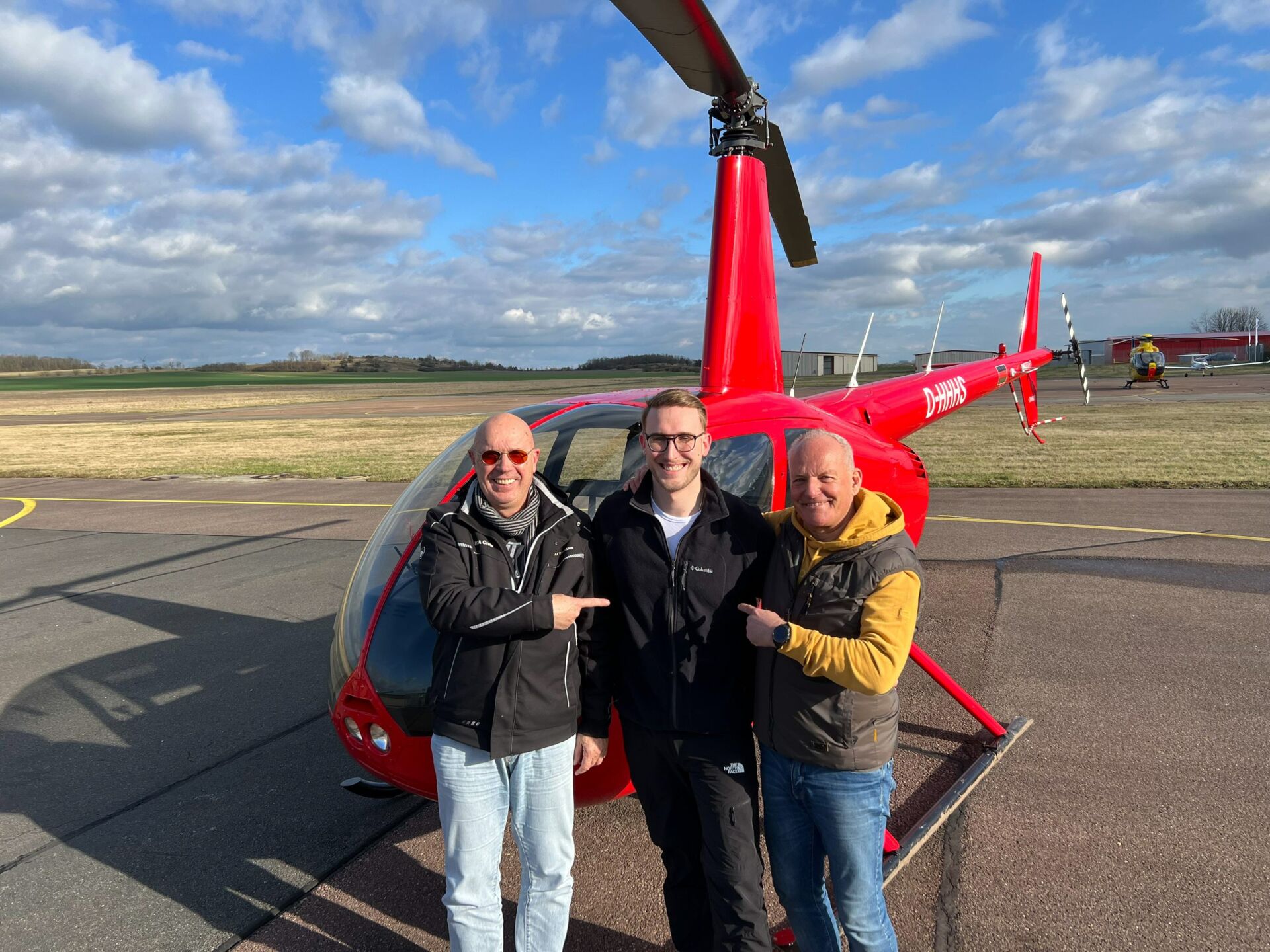 Drei Männer stehen vor einem roten Helikopter auf einem Fluglandeplatz.
